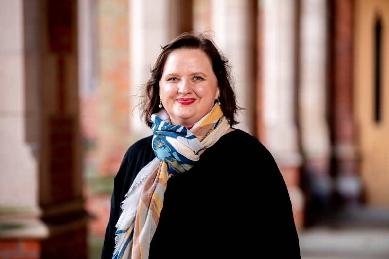 smiling woman with shoulder length brown hair, wearing a black top and multi-coloured scarf, with a red brick wall in background